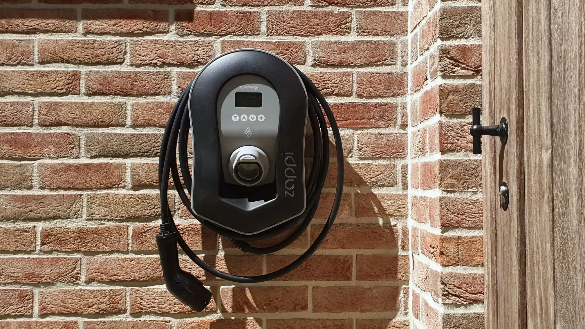 Laadpaal met opgerolde kabel aan een bakstenen gevel, naast een houten deur